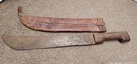 Machete with attached sheath lying on carpeted surface, showing full length of blade and sheath