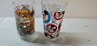 Photo showing two tall clear glass collectible drinking glasses on a white surface with colorful cartoon designs featuring vintage and Mickey Mouse themes.
