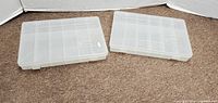 Two clear plastic Plano storage boxes seen from above on a carpeted floor showing the closed lids with visible grid compartments underneath.