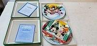 Two Schmidt Disney collectible plates side by side with certificates of authenticity inside green boxes