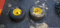 Two yellow riding mower tractor wheels with rims placed on asphalt, showing tread and mounting holes.