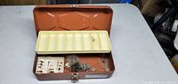 Open brown metal tackle box showing yellow plastic tray with compartments containing fishing hooks and lures.