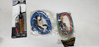 Photo shows three items: one electric tester in branded packaging on the left, two sealed packs of bungee cords to the right with blue and red/green cords.
