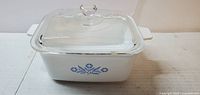 White rectangular CorningWare baking dish with clear glass lid and blue cornflower pattern on the side, shown from an angle.