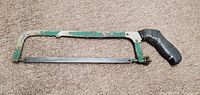 Metal hacksaw with green painted metal frame and black tape wrapped around the handle. Blade secured with wingnuts. Shows signs of wear and paint chipping.