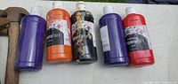 Photo of five large paint bottles: purple, orange, black, purple, red tempera paints, arranged on a table.