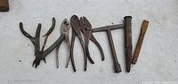 Photo showing eight small vintage hand tools on a white surface: wire cutter pliers, cutting pliers, two pairs of adjustable pliers, T-handle wrench, rusty hammer head without handle, wooden-handled metal tool.