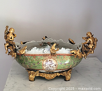 Frontal view of oval porcelain bowl with green ground, gilt cherub handles and ornate gilt-metal base.