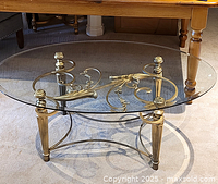 Metal and glass coffee table with an oval clear glass top and ornate gold-finished metal base with decorative scrollwork and finials visible.