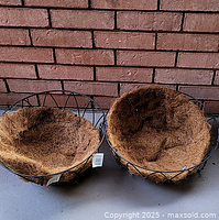 Pair of black metal hanging baskets with coir liners inside, shown from above resting on concrete floor, brick wall background.