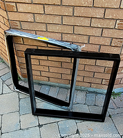 Pair of square-shaped black metal table legs resting upright against a brick wall outdoors on stone pavement.