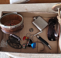 Various small decorative items in a cardboard box including a vintage iron, glass lampshade, metal flask, wooden drum, and small figurines.