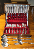 Full set of silver plated flatware displayed in an open wooden box with red velvet lining, showing multiple forks, spoons, and knives securely held in slots.