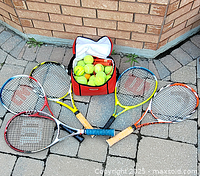 Five tennis rackets arranged on a patio surface with a soft sports bag filled with tennis balls in the center. Rackets are Wilson brand.