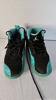 Front view of the black and turquoise Air Jordan 12 Retro kids sneakers showing the shape, laces, and logos.