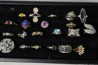 Full view of approximately 18 assorted costume rings arranged in a black tray with various colorful stones and metal finishes.