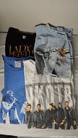 Four folded concert T-shirts piled showing Lady Zephyr, Kenny Chesney, UKOTB, and a blue shirt with singer graphic.