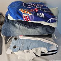 Stack of folded clothing including a New York Giants champion T-shirt on top, dark and light denim jeans, and folded shirts with tags.