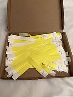 Open cardboard box filled with many yellow and white event admission paper bracelets stacked loosely inside.