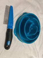 Both items shown together on white fabric background: blue slow feed dog bowl and Wahl branded fine tooth comb