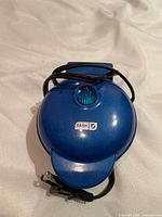 Closed blue mini waffle maker with electric cord coiled on top, showing brand logo 'Dash'.