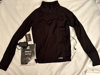 Black heated jacket / thermal top laid flat on white surface with power adapter and RPHA 1.0 battery pack, showing front side and zippered chest pocket.