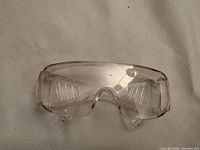 Photo shows one pair of clear transparent protective safety glasses with side shields lying flat on a beige fabric surface.