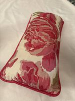 Rectangular lumbar pillow with floral rose pattern in red and pink shades, edged with red piping, lying on a white surface.