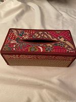 Top view of rectangular tissue box cover with red silk blend fabric embroidered with floral and abstract patterns and an oval opening for tissues.