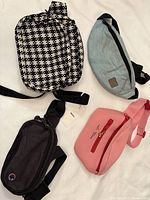 Four fanny packs laid out on a white surface: Baggu patterned black and white, Champion gray, Tinyat black, and pink Marc Jacobs.