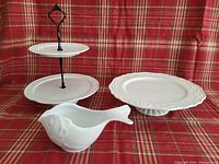 Photo showing 2-tier dessert stand with black metal rod and white ceramic trays, filigree pedestal plate with ornate edges, and fish-shaped gravy boat on red plaid background.
