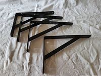 Four black metal shelf brackets arranged on a white cloth surface, showing the triangular support design and mounting holes.