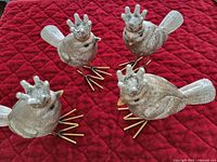 Four gold-colored bird figurines with crowns arranged on a red quilted cloth, all facing slightly different directions showing their glittery textured finish and metallic legs.