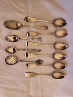 Summary photo of all 10 antique sterling silver teaspoons showing various handle designs, including a slotted spoon and a spoon with decorative emblem.