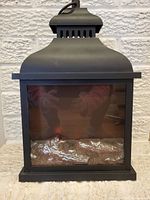 Front view of the black metal fireplace lantern with glass panel and faux logs inside. Lantern is not lit.
