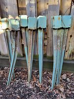 Seven light blue metal fence post spikes, leaned against a wooden fence, showing length, pointed end, and surface rust