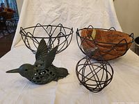 Photo shows two iron hanging baskets; left basket without liner and right basket with fiber liner. Also visible are green iron bird figurine and metal orb with small bird on top.