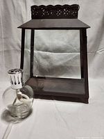 Front view of metal lantern with clear glass panels and decorative cutout top, alongside original Lampe Berger Paris lamp with clear glass body and silver-tone top.