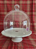 White ceramic pedestal cake plate with scalloped edge and clear glass dome cover set on a red plaid background.