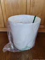 Large white planter in unused condition with plastic liner visible inside, placed on wood floor against cabinet backdrop.