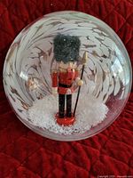 Full view of glass nutcracker snowglobe showing the nutcracker figurine inside standing on red base with white artificial snow flakes.