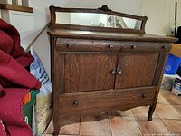 Front view of solid oak sideboard buffet with mirror on top, showing two top drawers, middle cabinet with double doors, and one bottom drawer. The wood has a dark finish and visible wear marks on top surface.