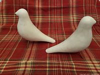 Pair of solid white marble dove sculptures shown side by side on a red plaid fabric background, profile view