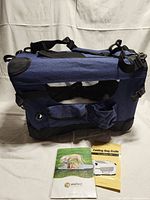 Photo showing full side view of blue folding dog crate with mesh panel, pockets, and shoulder strap, along with two instruction booklets on ground in front.
