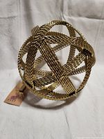 View of woven brass sphere from front, showing intricate woven brass detail and tag indicating brand new condition.