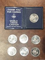 Overview of seven coins: six Windsor Anniversary Dollars arranged below one World Vision coin in holder