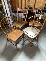 Photo of four different antique solid wood chairs grouped together on concrete floor in wooden framing environment. Shows details and style variation of chairs.