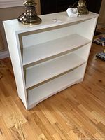 White painted wooden bookcase showing full front with three shelves, placed on wooden floor with items on top