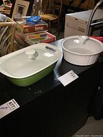 Two CorningWare bakeware dishes with lids on black surface with board games in background.