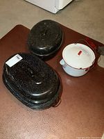 Two large black speckled enamel roasting pans with lids and one white enamel pot with a red handle on a wire bail and a pour spout, showing general wear and surface chipping.